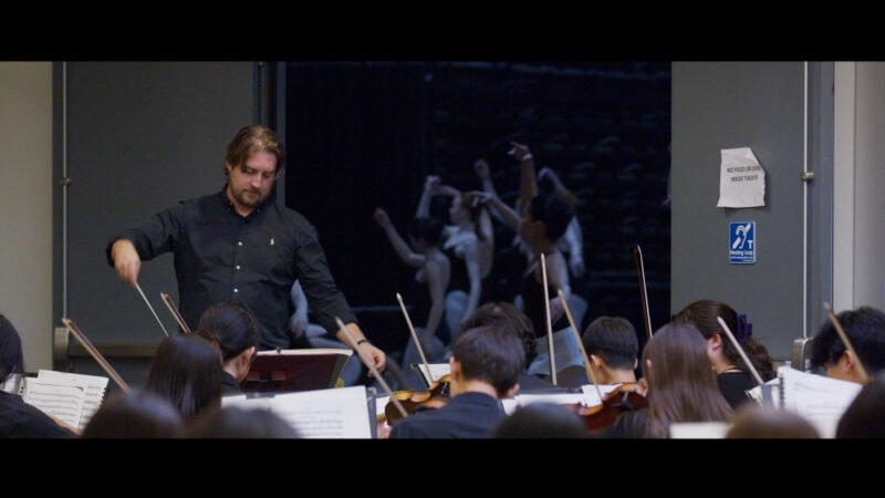 Orchestra conductor in black rehearsing with a youth orchestra and ballet dancers in the background.